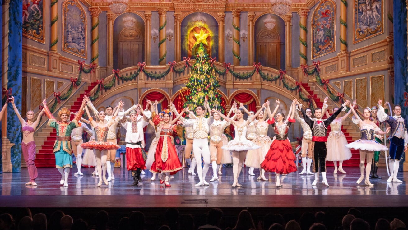 Ballet performers in colorful costumes hold hands and bow on an ornate stage decorated with a Christmas tree and lights.