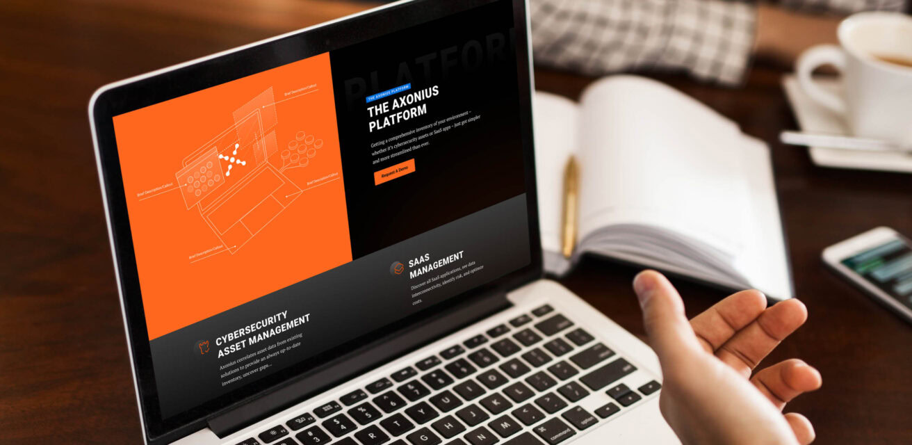 A person is using a laptop displaying a website for a platform focused on cybersecurity asset management and SaaS management, with an orange and black design theme.