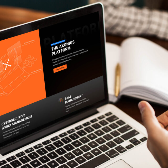 A person is using a laptop displaying a website for a platform focused on cybersecurity asset management and SaaS management, with an orange and black design theme.